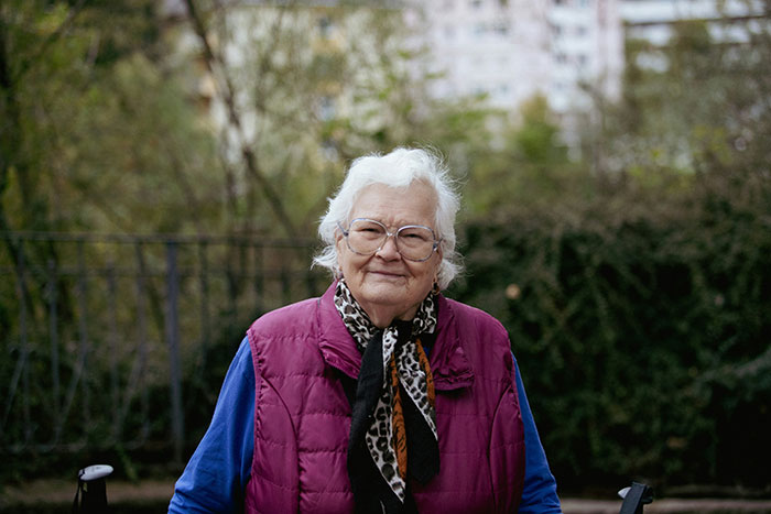 Elderly woman outdoors wearing glasses and a purple vest, representing family care and refusal to take responsibility.