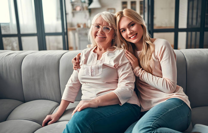 Pregnant woman sitting with her supportive mother at home, smiling and embracing on a gray sofa in a bright living room.