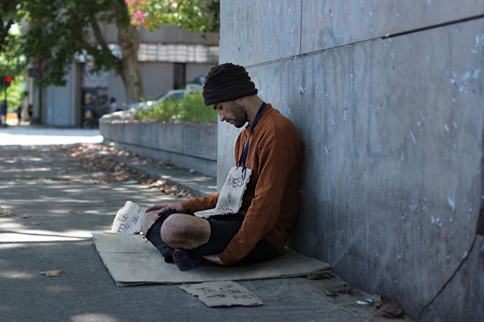 Young man sitting outside on a sidewalk, appearing homeless after parents kicked out teen, wearing a beanie and brown jacket.