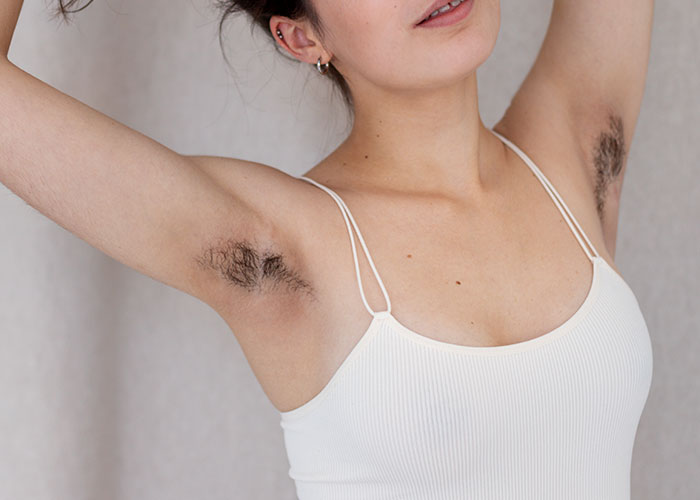 Woman showing hairy armpits in a white tank top, highlighting bridal guests feeling uncomfortable with body hair.