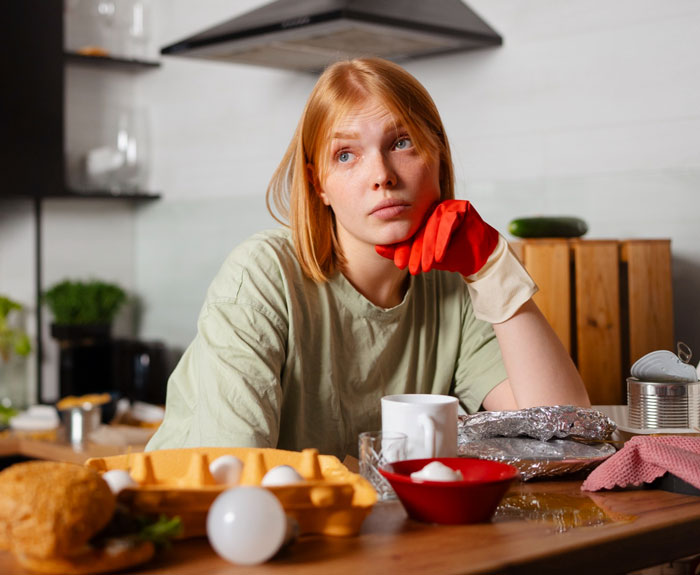Young woman in kitchen looking frustrated while wearing a red glove, surrounded by food and cooking items reflecting picky-eater MIL issues